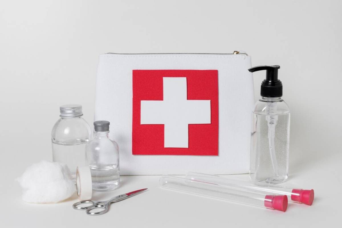 A first aid kit with a red cross symbol, flanked by bottles, cotton, scissors, and test tubes on a light background.