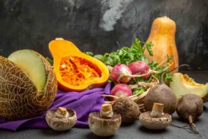 A colorful array of fresh vegetables, including melon, pumpkin, mushrooms, radishes, and herbs, set on a purple fabric backdrop.