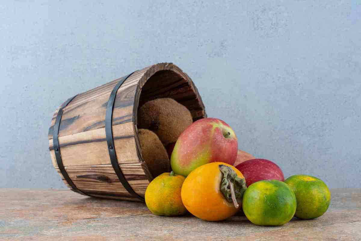 A wooden bucket tipped over, spilling various fruits including apples, kiwis, and citrus, set against a light gray background.