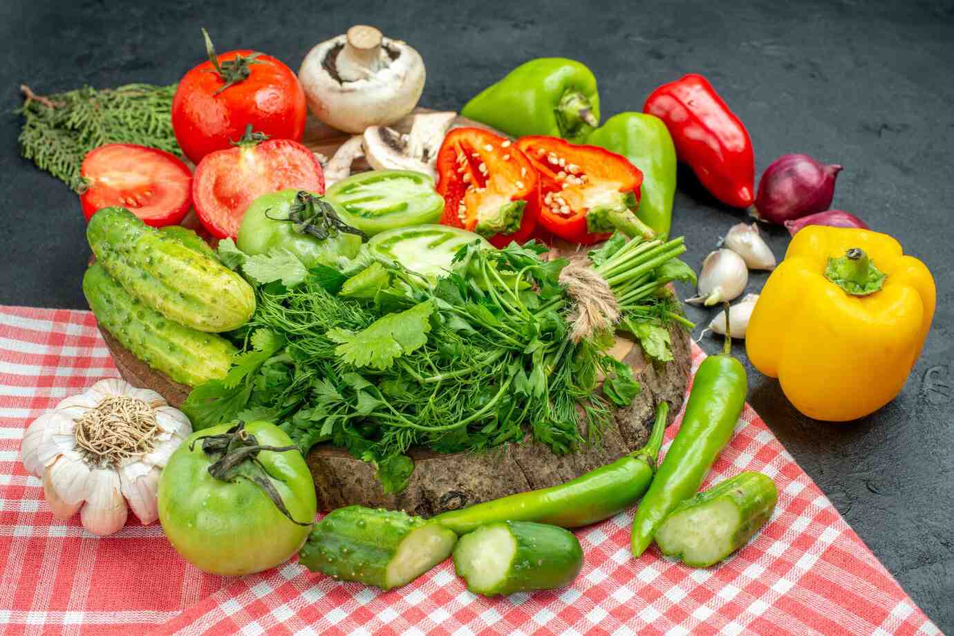 A vibrant assortment of fresh vegetables including tomatoes, peppers, cucumbers, mushrooms, and herbs on a wooden board with a checkered cloth.